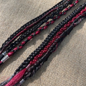 Può includere: Due trecce di extension per capelli nere e bordeaux. Le trecce sono intrecciate insieme con un motivo semplice.