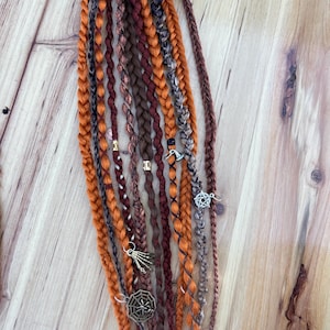 Peut inclure: Un ensemble d'extensions de cheveux synthétiques brunes, orange et noires avec des breloques. Les breloques comprennent une toile d'araignée, une main de squelette et un pentagramme.