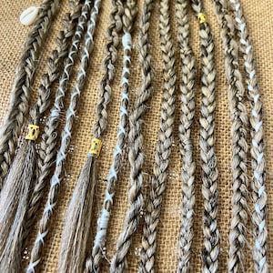 Peut inclure: Une collection de tresses brunes et blondes avec des accents de ficelle blanche et des perles dorées. Les tresses sont disposées sur un fond de toile de jute brune.