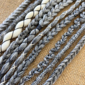 Op de afbeelding: Een set grijze en witte synthetische haarverlengingen, gevlochten en aan elkaar gebonden met zwart garen. De verlengingen zijn in een rij gerangschikt, met de witte verlengingen bovenaan en de grijze verlengingen onderaan.