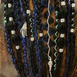 May include: Braided black and blue dreadlocks with silver accents and charms. The dreadlocks are adorned with silver beads, a hand charm, and a skull charm on a chain. Teal and blue strands are woven throughout the braids.