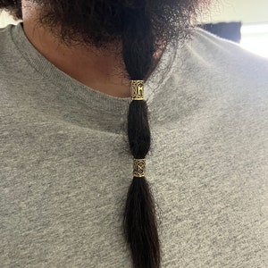 May include: A man with a long braid of hair tied with three gold beads. The beads have intricate designs.