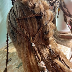 May include: A close-up of a mannequin head with long, light brown hair styled with multiple braids. The braids are interwoven and adorned with small silver accents and white thread. The hairstyle is intricate and detailed.