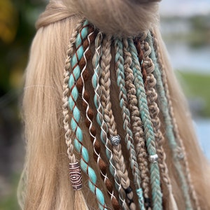 May include: Close-up of blonde hair styled with multiple braids and decorative accents. Braids feature shades of brown, teal, and white, with small beads and charms. The hair is styled in a half-up, half-down style.