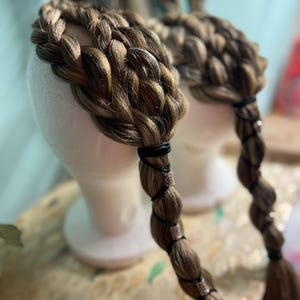 May include: Two braided wigs with brown and blonde hair are displayed on white mannequin heads. The braids are secured with black bands and adorned with silver-colored metal accents. The wigs are designed to resemble a crown.