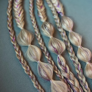 Könnte beinhalten: Nahaufnahme von mehreren blonden Zöpfen mit lila Glitzerakzenten. Jeder Zopf ist mit einer Reihe von geknoteten Abschnitten versehen, wodurch ein einzigartiges und dekoratives Aussehen entsteht. Die Zöpfe sind auf einer grauen Oberfläche angeordnet.