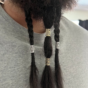 May include: Close-up of three braided sections of dark hair, each adorned with metallic hair beads. The beads feature intricate, engraved designs. The hair is styled with a gray shirt in the background.