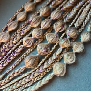 Könnte beinhalten: Nahaufnahme von mehreren blonden geflochtenen Haarverlängerungen mit dekorativen Knoten. Die Zöpfe haben Strähnen aus lila und schillerndem Glitzer. Die Verlängerungen sind diagonal vor einem gedämpften blau-grünen Hintergrund angeordnet.