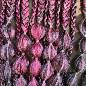 Könnte beinhalten: Nahaufnahme von mehreren Zöpfen in Rosa-, Schwarz- und Weißtönen. Die Zöpfe sind in vertikalen Reihen angeordnet, wobei jeder Zopf eine Reihe von knolligen, geknoteten Abschnitten aufweist. Der Gesamteffekt ist eine strukturierte und farbenfrohe Darstellung, geeignet für das Haarstyling.