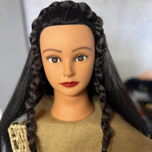 May include: A mannequin head with long, dark brown hair styled in two braids. The braids are secured with black elastic bands and the hair is parted in the middle. The mannequin has a neutral expression with brown eyes and red lips.