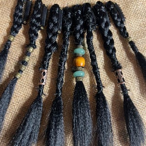 May include: Black braided hair extensions with metal clips and decorative beads. The extensions are arranged in a fan shape, with each braid ending in a loose, textured fringe. The beads are in shades of gold, copper, and turquoise.
