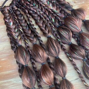 Könnte beinhalten: Geflochtene braune Haarverlängerungen mit irisierenden Strähnen. Die Extensions sind mit mehreren geflochtenen Haarpartien versehen, die jeweils mit einem schwarzen Gummiband befestigt sind. Das Haar liegt auf einer Holzoberfläche.