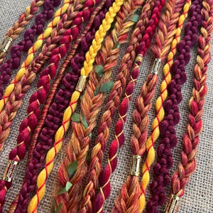May include: A set of braided hair extensions in shades of burgundy, red, orange, and yellow. The extensions are decorated with gold beads.