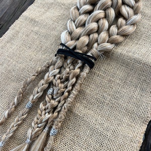 May include: A set of four blonde braids with white highlights, tied together with a black elastic band. The braids are decorated with silver beads.