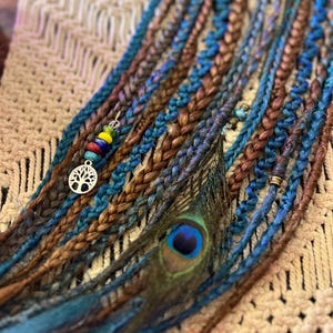 May include: A set of synthetic hair extensions featuring a blend of brown and blue braided strands. The accessory is decorated with a silver tree of life charm, small colorful beads, and a single peacock feather attached to the braids.