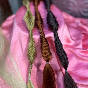 May include: Three braided hair extensions in blonde, brown, and black colours. The extensions are displayed on a pink satin surface.