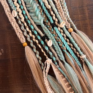 Peut inclure: Gros plan sur plusieurs extensions de cheveux tressées dans des tons marron, beige et turquoise. Les extensions sont ornées de coquillages, de perles en bois et de plumes. Certaines tresses sont enroulées de fil turquoise. Les extensions sont disposées sur une surface marron foncé.