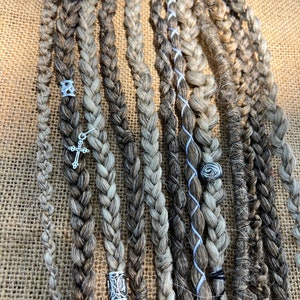 May include: Braided dreadlocks in shades of brown and gray, adorned with silver-colored beads and a cross charm. The dreads are arranged on a textured, tan surface, showcasing the intricate braiding and varied tones.