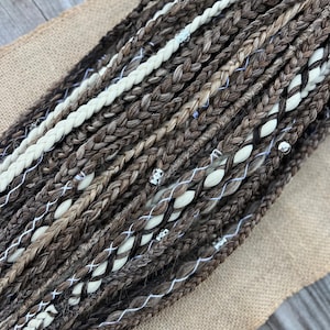 May include: A close-up of a bundle of brown, black, and white braided hair extensions. The extensions are arranged in a fan shape and have silver beads woven into them.