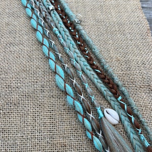 Peut inclure: Gros plan sur plusieurs extensions de cheveux tressées dans des tons marron, gris et turquoise. Certaines tresses sont ornées de motifs en point de croix blancs et de petits charms, dont un coquillage. Les extensions sont disposées sur une surface texturée et beige.