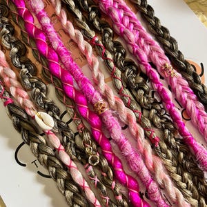 Peut inclure: Une collection d'extensions de cheveux synthétiques dans différentes nuances de brun, rose et blanc. Les extensions sont tressées et attachées avec un ruban rose. Certaines extensions ont de petits charmes décoratifs.