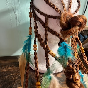 May include: Braided brown hair accessory with turquoise and tan feathers, wooden beads, and decorative accents. The hairpiece is styled on a white mannequin head, showcasing its intricate design and bohemian aesthetic.