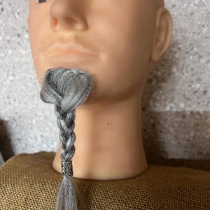 May include: A mannequin head with a gray braided beard. The beard is styled with a silver-colored metal bead at the end of the braid. The mannequin is on a burlap surface.