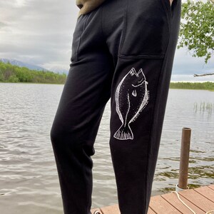 May include: Black sweatpants with a white graphic of a fish on the left leg. The fish is outlined in white and has a dotted pattern. The text "Prince William Sound" is written on the fish.