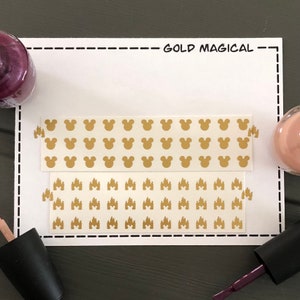 May include: Two sheets of gold nail decals. One sheet features a repeating pattern of Mickey Mouse heads. The other sheet features a repeating pattern of the Cinderella Castle silhouette. The text "GOLD MAGICAL" is written on a white card.
