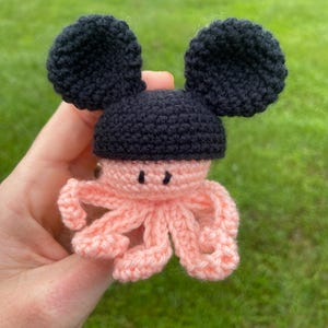 May include: A handmade crochet octopus toy with black ears and head, and a pink body and tentacles. The toy is held in a hand against a blurred green background.