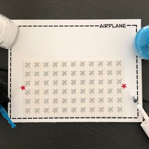 May include: A sheet of white sticker paper with a repeating pattern of gray airplane silhouettes. The sheet is surrounded by a black dashed line border. The word "AIRPLANE" is printed in black at the top of the sheet. Two red stars are placed on either side of the sheet.