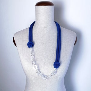 Op de afbeelding: Een blauwe en transparante ketting met een geknoopt touwontwerp en een transparante ketting. De ketting wordt getoond op een wit mannequin.