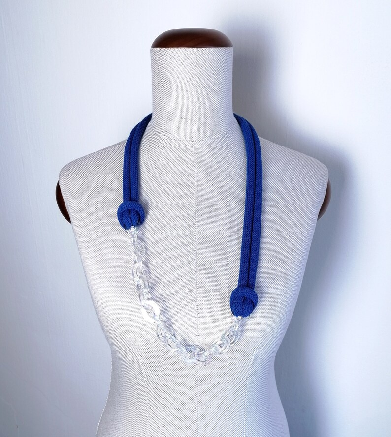 Op de afbeelding: Een blauwe en transparante ketting met een geknoopt touwontwerp en een transparante ketting. De ketting wordt getoond op een wit mannequin.