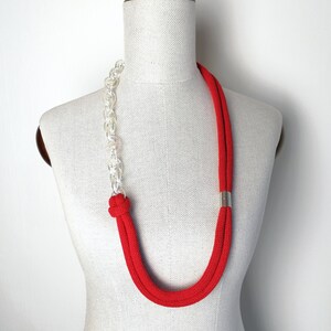 Op de afbeelding: Een rode en doorzichtige plastic ketting met een zilveren accent. De ketting is op een wit mannequin.