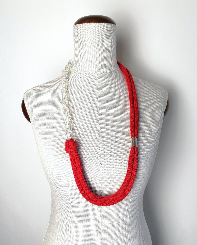 Op de afbeelding: Een rode en doorzichtige plastic ketting met een zilveren accent. De ketting is op een wit mannequin.