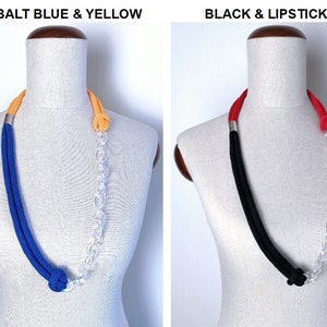 Op de afbeelding: Twee kettingen met een transparante plastic ketting en twee stoffen koorden. De eerste ketting heeft een kobaltblauw koord en een geel koord. De tweede ketting heeft een zwart koord en een lipstickrood koord.