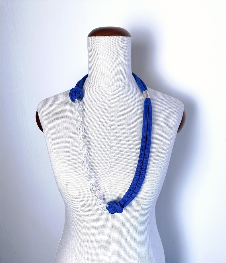 Op de afbeelding: Een blauwe en transparante plastic ketting met een zilveren accent. De ketting wordt getoond op een wit mannequin.
