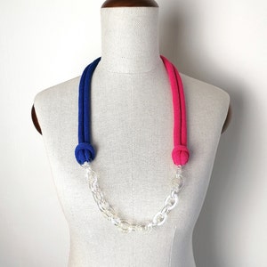 Op de afbeelding: Een blauwe en roze touwketting met een transparante plastic ketting. De ketting is aan de uiteinden geknoopt en wordt getoond op een wit mannequin.