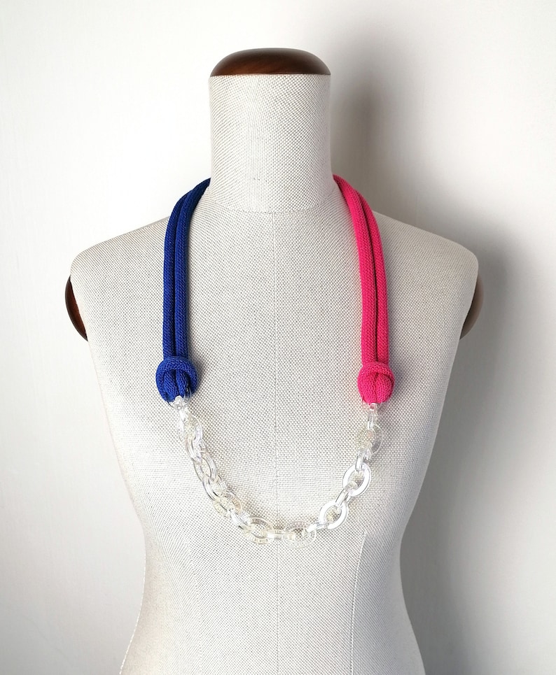Op de afbeelding: Een blauwe en roze touwketting met een transparante plastic ketting. De ketting is aan de uiteinden geknoopt en wordt getoond op een wit mannequin.