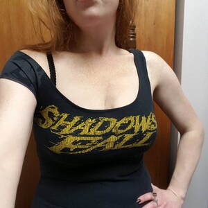 Puede incluir: Un top negro de tirantes con un gráfico dorado que dice "Shadows Fall".