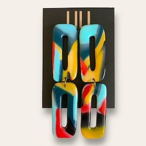 May include: A pair of colorful, abstract resin earrings. The earrings are rectangular in shape and have a blue, yellow, red, and black color scheme.
