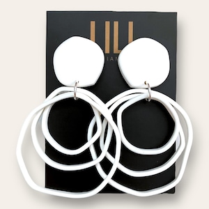 Puede incluir: Pendientes de cerámica blanca con una forma de círculo grande e irregular y varios círculos más pequeños superpuestos. Los pendientes se muestran sobre un fondo negro con la palabra "LILA" en letras doradas.