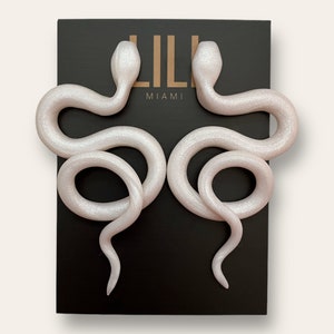 May include: A pair of white snake-shaped earrings with a sparkly finish. The earrings are on a black background with the text "LILI MIAMI" printed on it.