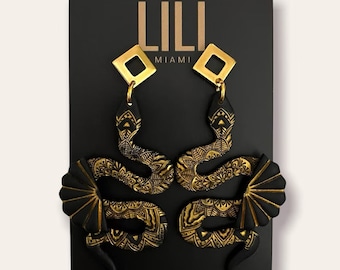 Puede incluir: Un par de pendientes de serpiente negros y dorados con un diseño de mandala. Los pendientes están en una tarjeta negra con el texto "LILI MIAMI" en letras doradas.