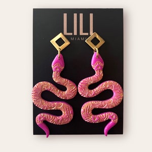 May include: A pair of pink snake earrings with gold accents. The snakes are detailed with a floral pattern and have gold square charms at the top. The earrings are displayed on a black background with the text "LILI MIAMI".