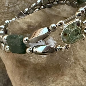 May include: Silver bracelet featuring green square and oval beads, and a turtle charm. The bracelet is composed of small, round beads and is shown on a textured, neutral surface. The turtle charm has a green, patterned centre.