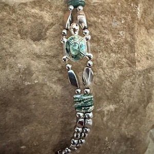 May include: A silver bracelet with a turtle pendant, featuring a green and blue shell. The bracelet includes silver beads, green stone beads, and leaf-shaped silver accents. The bracelet is displayed on a textured stone surface.