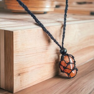 Può includere: Una collana con un ciondolo in pietra di corniola, un cordoncino nero e un design a nodo macramé. Il ciondolo è una pietra naturale di colore marrone-arancione.