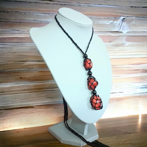 May include: A long necklace with three orange gemstone pendants wrapped in black macrame. The necklace is displayed on a white mannequin.