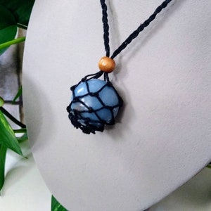 以下が含まれることがあります： 水色の宝石ペンダントネックレス。宝石は黒いマクラメネットに包まれ、黒いコードから吊り下げられています。木製のビーズがコードをアクセントにしています。ネックレスは白いバストに飾られています。